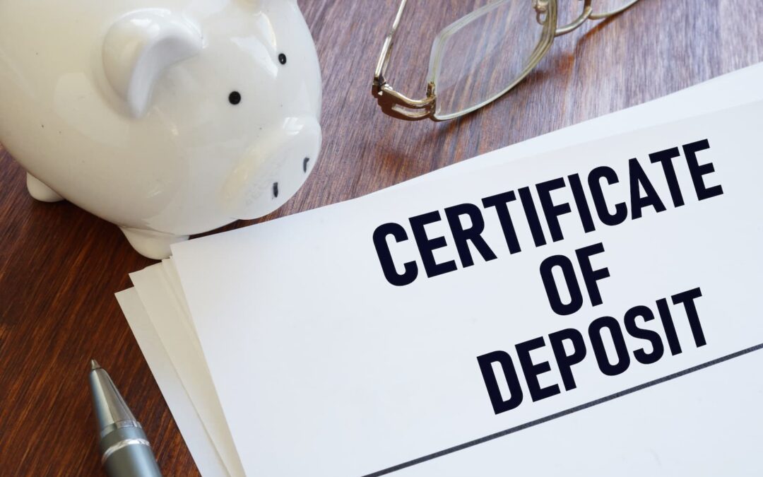 An overhead photo of a wooden desk with a stack of papers, on which a document is visible with the bold, large text "CERTIFICATE OF DEPOSIT." A white piggy bank and a pair of glasses are in the top left, and a pen is at the bottom left. The objects are arranged to suggest financial planning or savings.