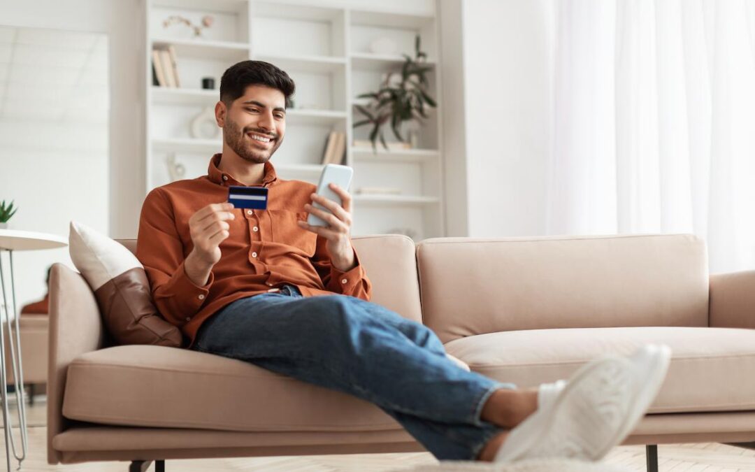 A smiling young man is relaxed on a beige couch in a brightly lit living room, holding a credit card in one hand and a smartphone in the other. He is looking at the phone, likely making an online purchase or banking transaction. He is wearing an orange button-down shirt, blue jeans, and white slippers or sneakers, with his feet resting on a footstool. The background includes a white shelving unit with books and plants.