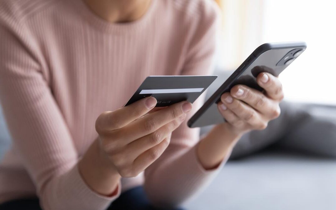 A close-up shot of a person's hands holding a black credit or debit card and a smartphone. The person appears to be performing an online transaction or entering card details into a banking app. They are wearing a light pink long-sleeved shirt. The background is blurred, suggesting an indoor setting like a home or office.