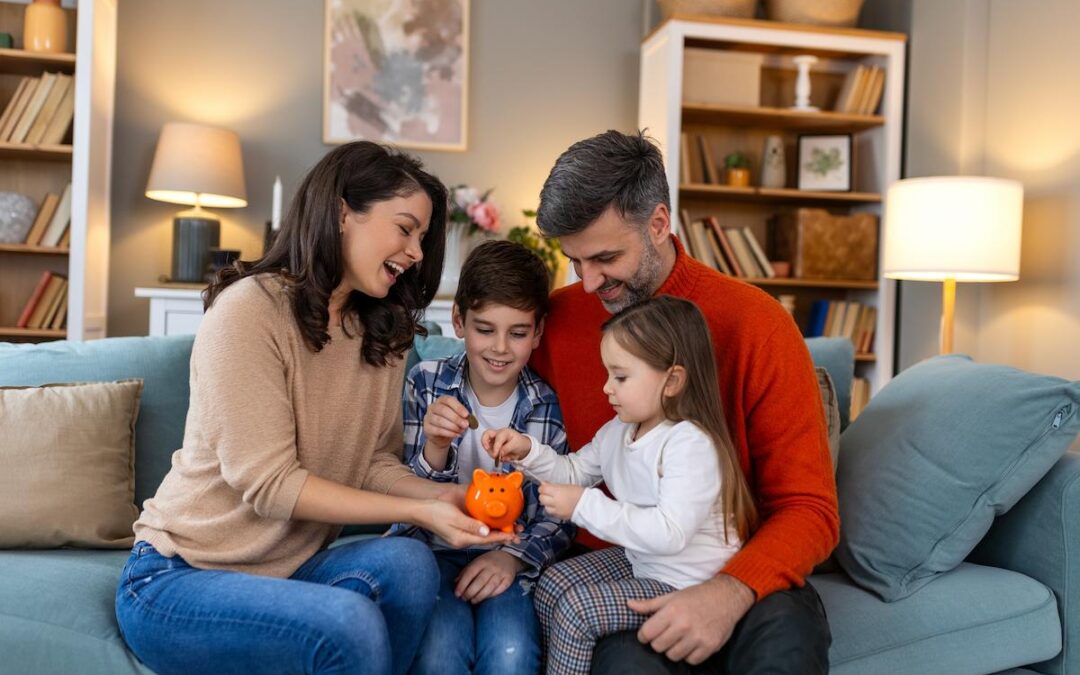 A cheerful family of four is sitting on a blue couch in their living room, focusing on a bright orange piggy bank. The mother and father are smiling as their two young children, a boy and a girl, insert coins into the bank. The girl is sitting on the father's lap, and the family appears to be teaching the children about saving money. The background includes bookshelves, lamps, and a framed picture, creating a cozy atmosphere.
